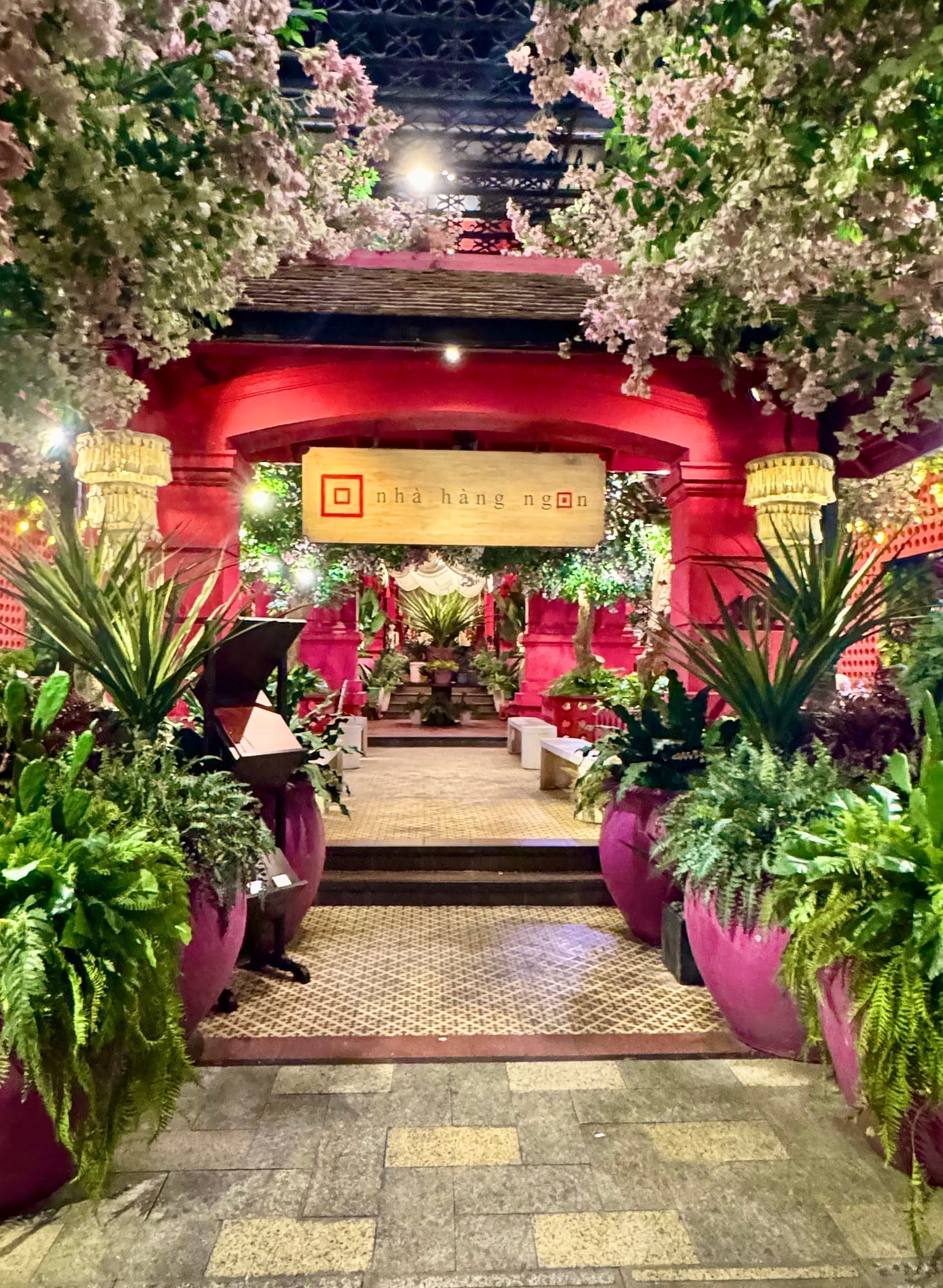Nhà Hàng Ngon entrance at night, decorated with hanging white and pink flowers and golden lanterns